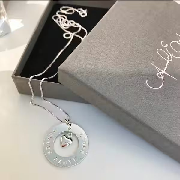 Silverhalsband med ringformad namnbricka och ett hjärta i mitten ligger utanför en smyckesask
